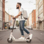 Mlad moški z nahrbtnikom vozi električni skiro po mestni ulici, obkrožen z večstanovanjskimi stavbami.